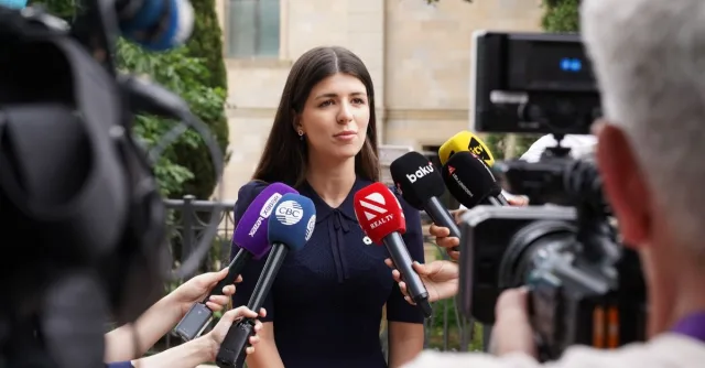 A spokesperson engages with media during an outdoor press interview surrounded by microphones.