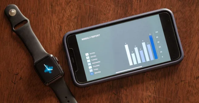 Close-up of a smartphone and smartwatch displaying a weekly report on a wooden table.