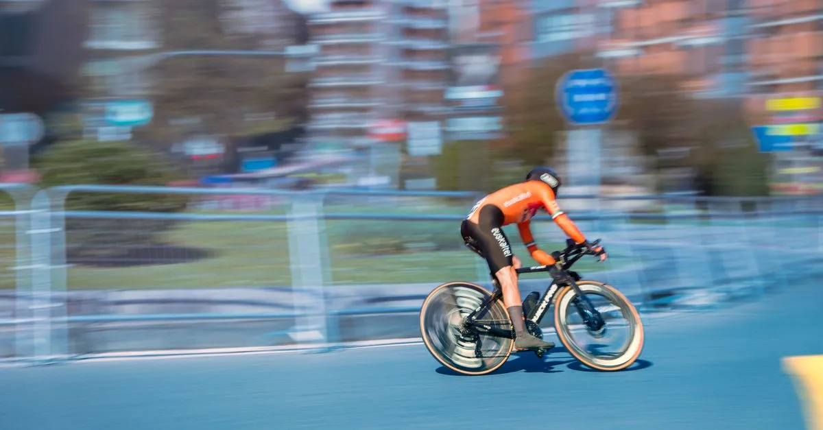 Blurry cyclist races through city streets wearing orange sportswear.