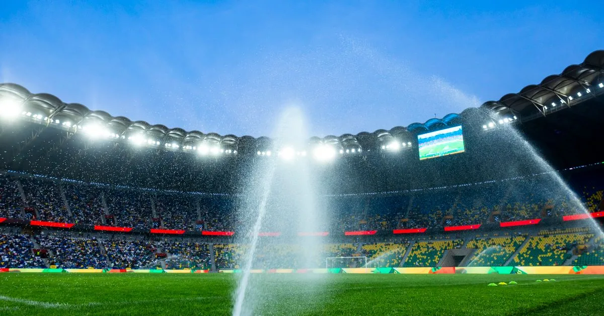 Nighttime view of a soccer stadium with sprinklers watering the field, enhancing the vibrant atmosphere.