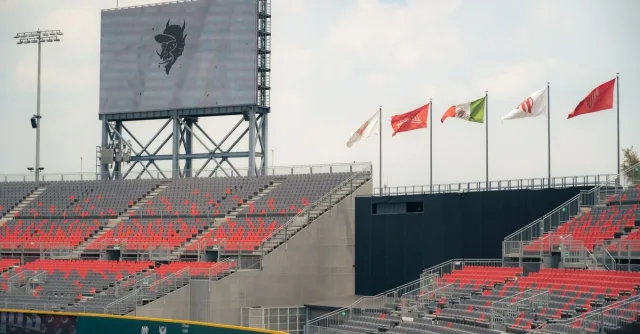 A modern stadium featuring red and gray seating and international flags, under a cloudy sky.