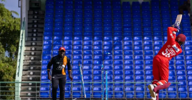 A cricket player in red batting during a match in an empty stadium with blue seats.