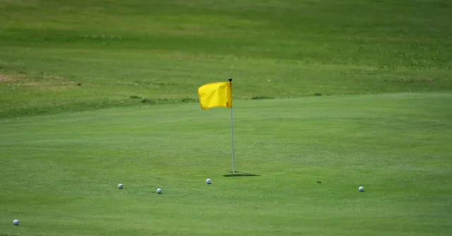 Golf course scene with yellow flag and golf balls on green under a clear sky.