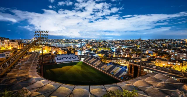 Aerial view of a brightly lit stadium surrounded by city buildings at dusk under a clear sky.