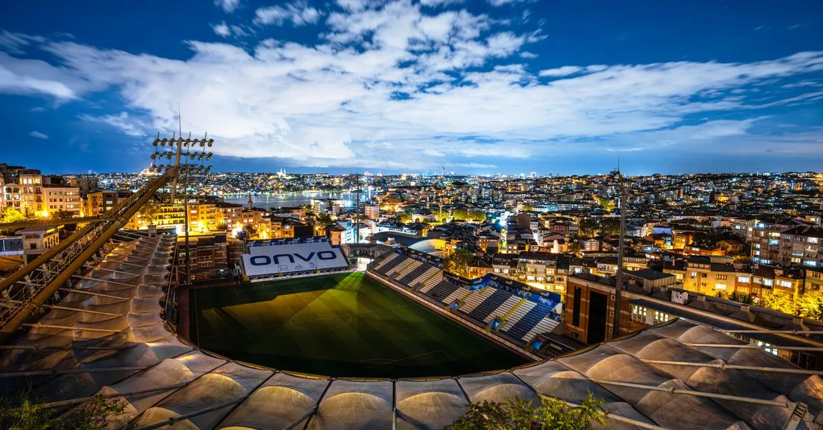 Aerial view of a brightly lit stadium surrounded by city buildings at dusk under a clear sky.