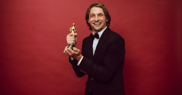 Smiling man in a tuxedo holding a trophy against a red background, celebrating success.