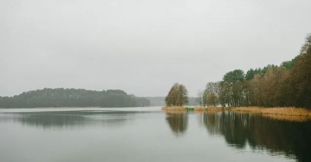 Tranquil lake scene in Poland surrounded by trees under a cloudy sky, reflecting serenity and peacefulness.