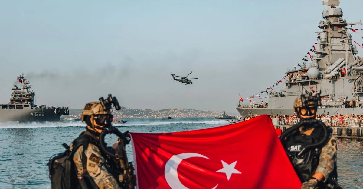 Military personnel holding a Turkish flag with naval ships and helicopter in the background.