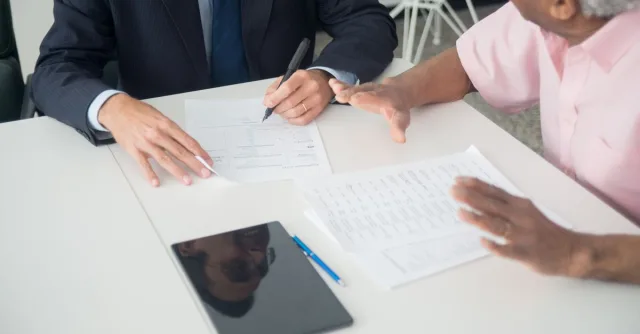 A business meeting between an elderly client and a consultant discussing documents at an office table.
