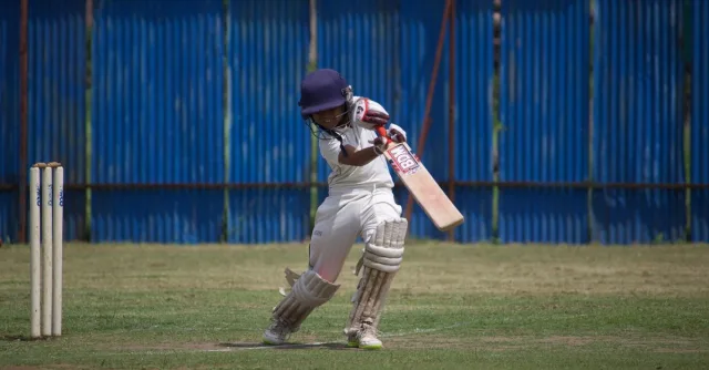 A young cricketer in action on a sunny day, playing on a grass field with a bat and wicket.