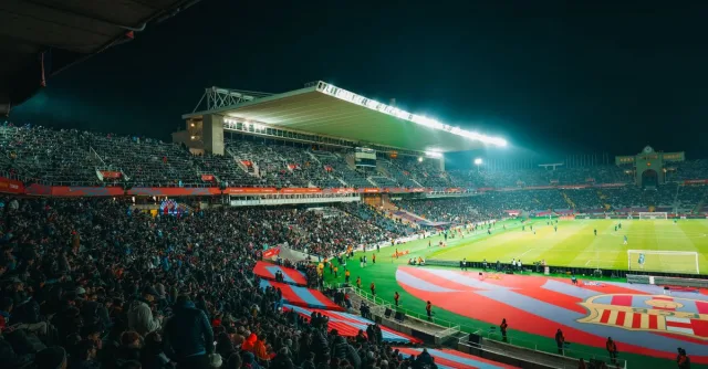 A crowded night-time football match in a brightly lit and bustling stadium, full of energy.