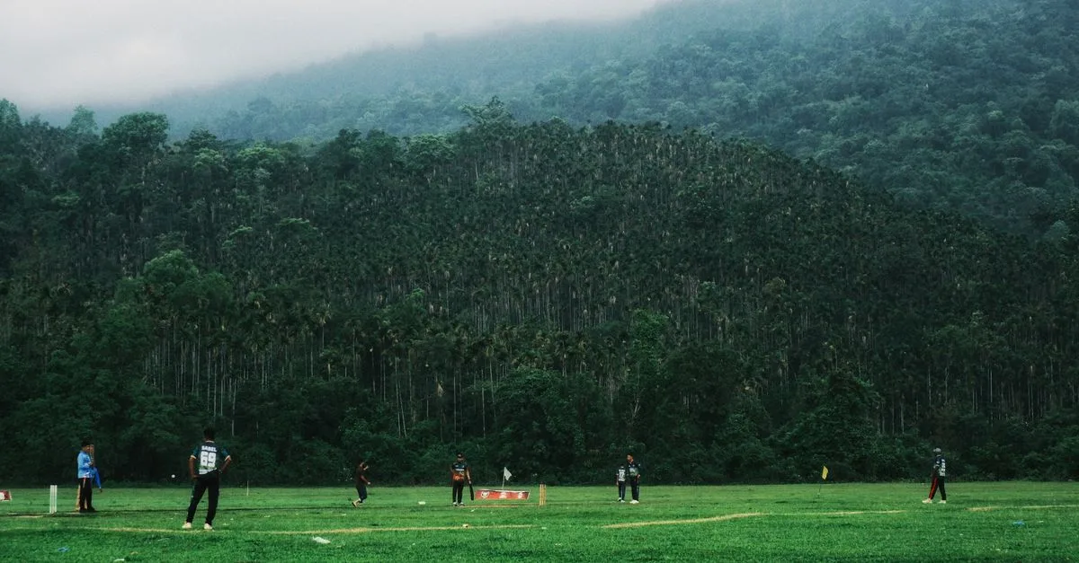 Cricketers playing in a lush green field surrounded by a forest in Sylhet, Bangladesh.