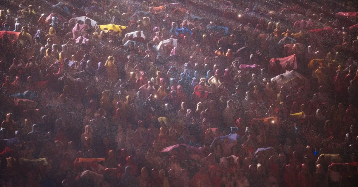 A large crowd gathers at night in the rain, illuminated by bright lights.