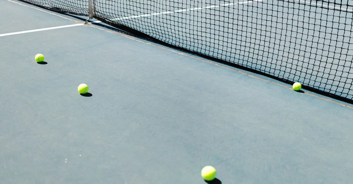 A tennis court featuring a net and several tennis balls lying on the ground, well-lit by sunlight.
