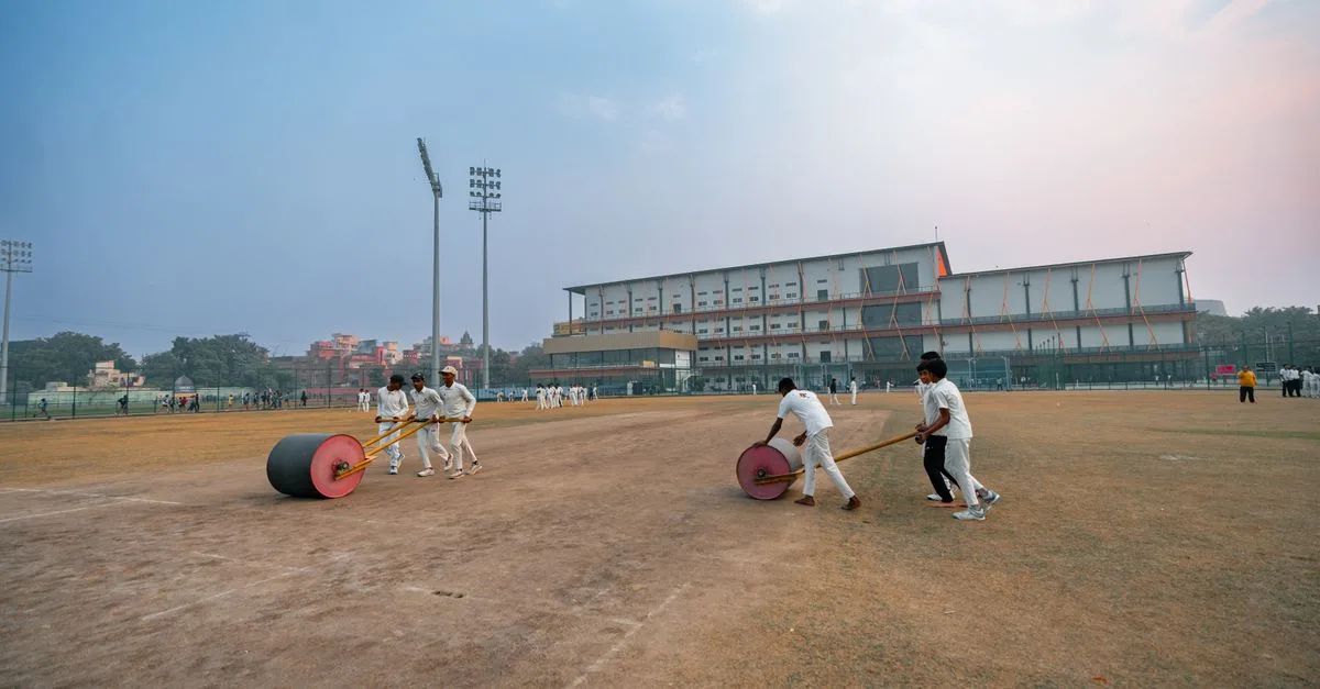 Groundskeepers preparing cricket field in Delhi, India with rollers at sunset.