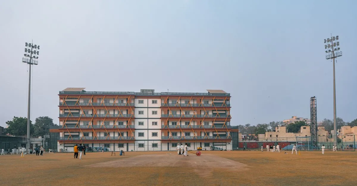 Daytime cricket match on a large sports field in Delhi, India with players and floodlights.