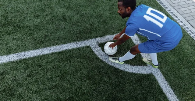 A soccer player in a blue jersey prepares to kick the ball from the corner on an artificial turf field.
