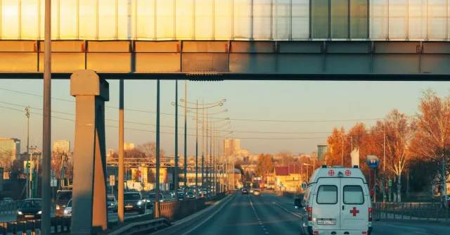 A serene view of an ambulance traveling on a highway in Perm, Russia during sunset, capturing autumn foliage and urban landscape.
