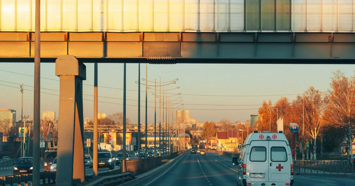 A serene view of an ambulance traveling on a highway in Perm, Russia during sunset, capturing autumn foliage and urban landscape.