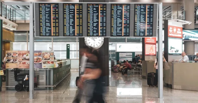 Blurred motion of travelers in a modern airport terminal with departure screens and a prominent clock.