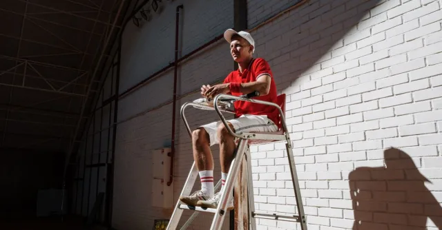 Tennis umpire sitting on a high chair against a white brick wall, creating dynamic shadows indoors.
