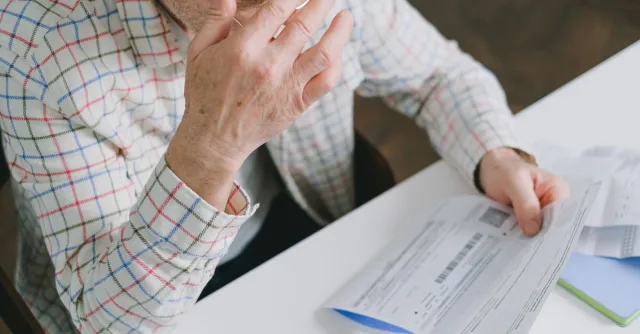 Elderly man in patterned shirt reading and holding bills at a home table, appearing focused.