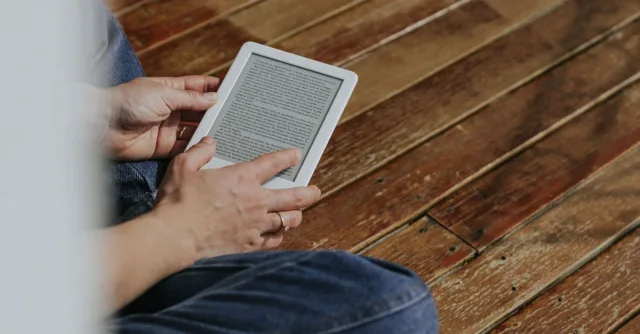 Close-up of hands holding an e-reader on a wooden floor, depicting a casual reading moment.