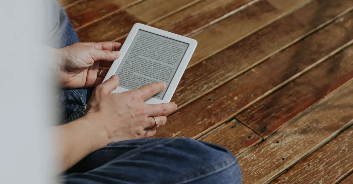Close-up of hands holding an e-reader on a wooden floor, depicting a casual reading moment.