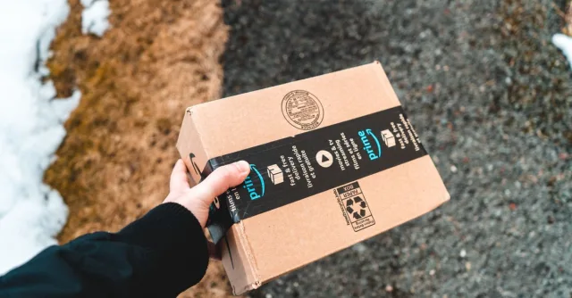 A person holding a cardboard Amazon Prime package on a snowy urban sidewalk.