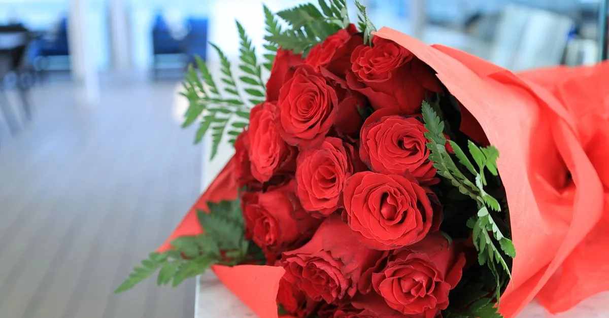 Close-up of a vivid red rose bouquet wrapped in orange paper with green leaves indoors.