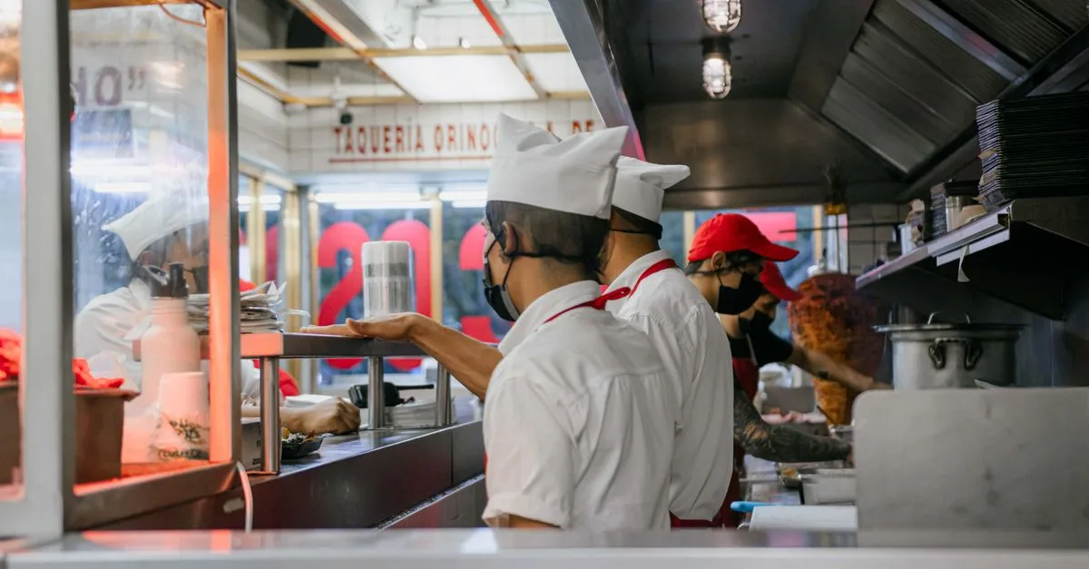 Culinary staff wearing uniforms and masks preparing meals in a bustling restaurant kitchen setting.