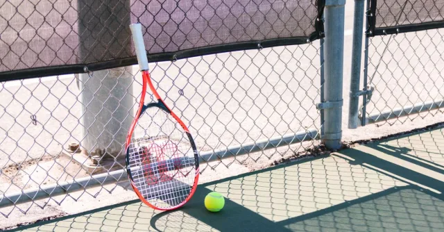 A tennis racket and ball rest on a court beside a chain-link fence in bright sunlight.