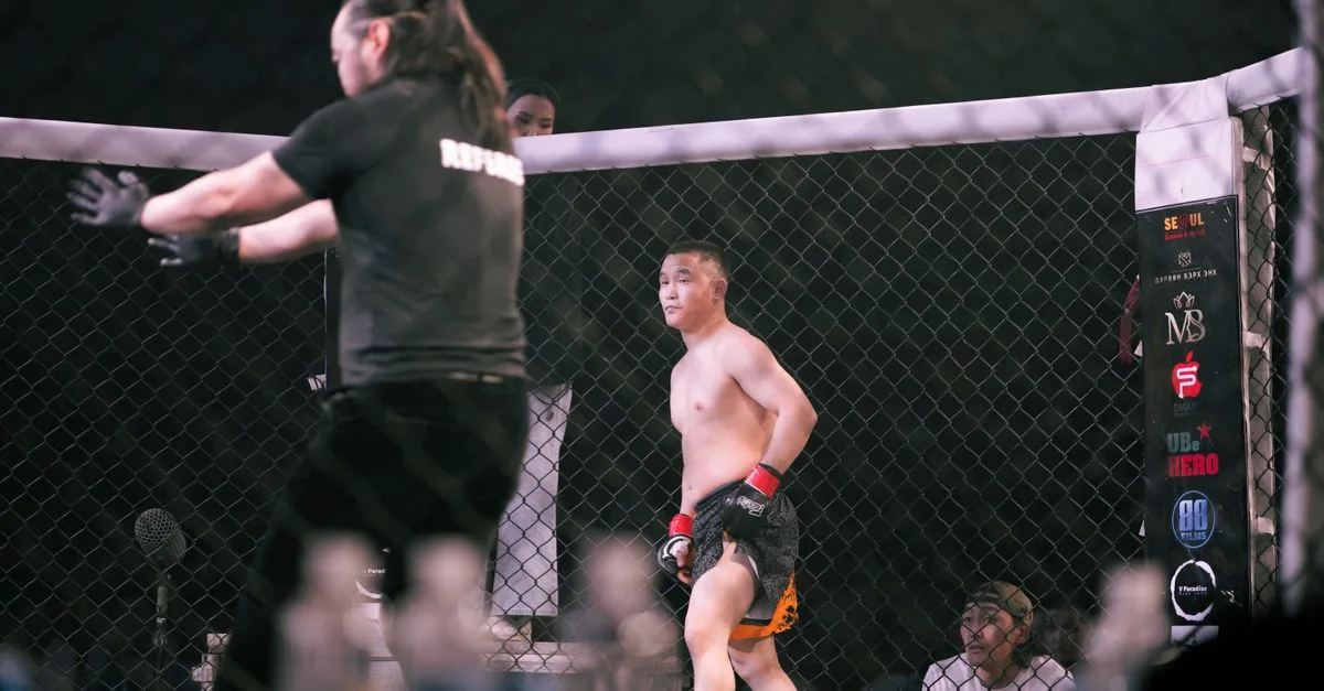 Mixed martial arts fight featuring two opponents and a referee in a caged arena.