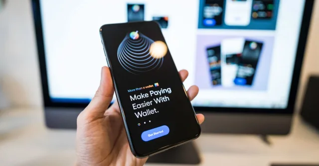 Hand holding smartphone displaying digital wallet app interface, blurred monitor in background.