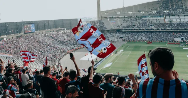Vibrant crowd cheers at a football match in a large stadium, waving team flags.
