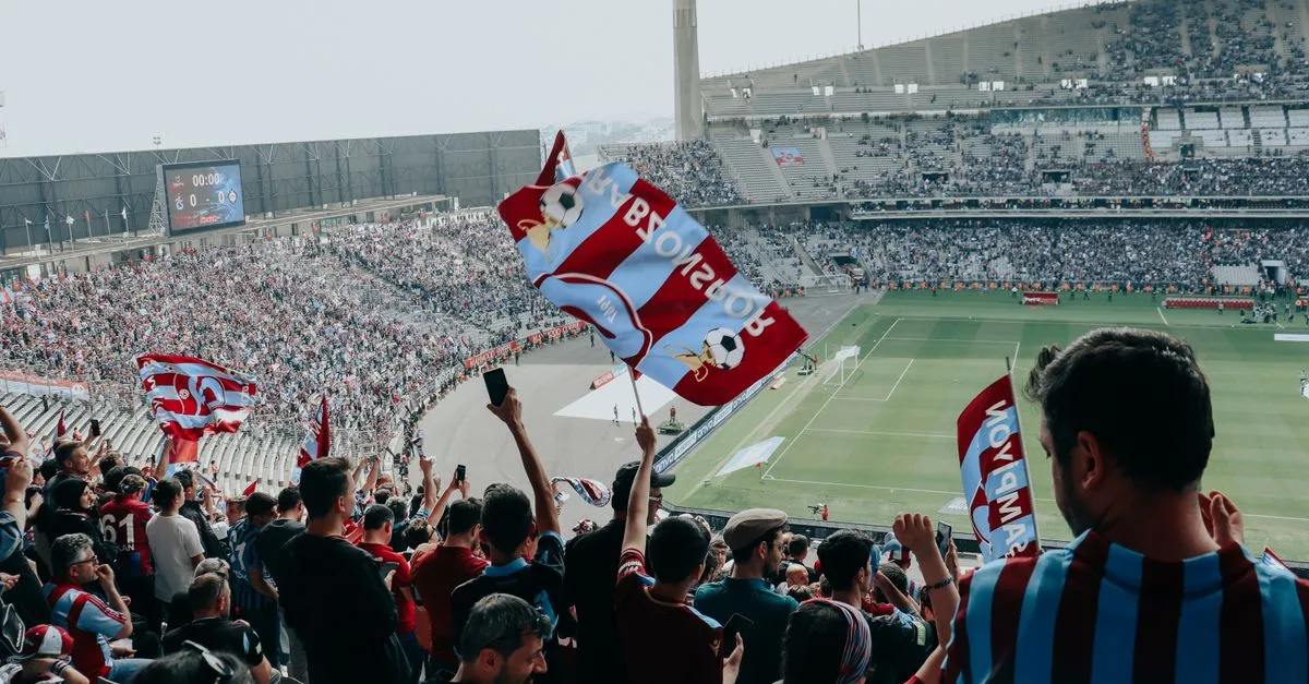 Vibrant crowd cheers at a football match in a large stadium, waving team flags.