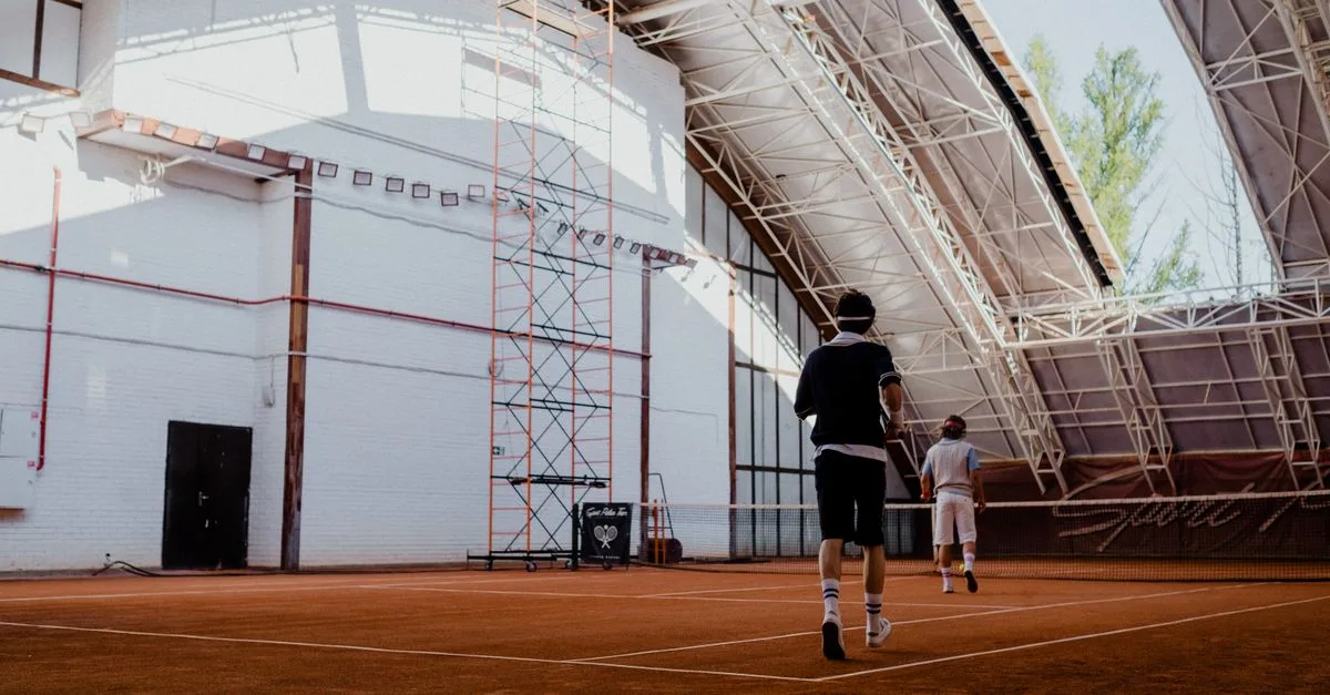 Two players engaging in a tennis match on an indoor clay court with natural lighting.