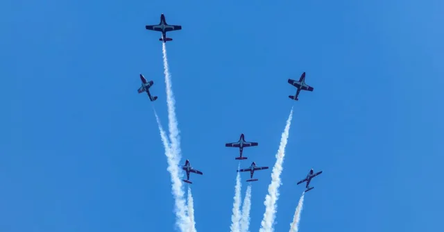 Six planes perform a synchronized aerial display with smoke trails in a clear blue sky.