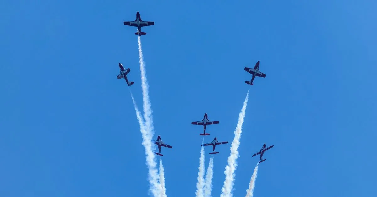 Six planes perform a synchronized aerial display with smoke trails in a clear blue sky.