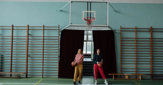 Two women holding basketballs in a gymnasium, standing by the hoop.