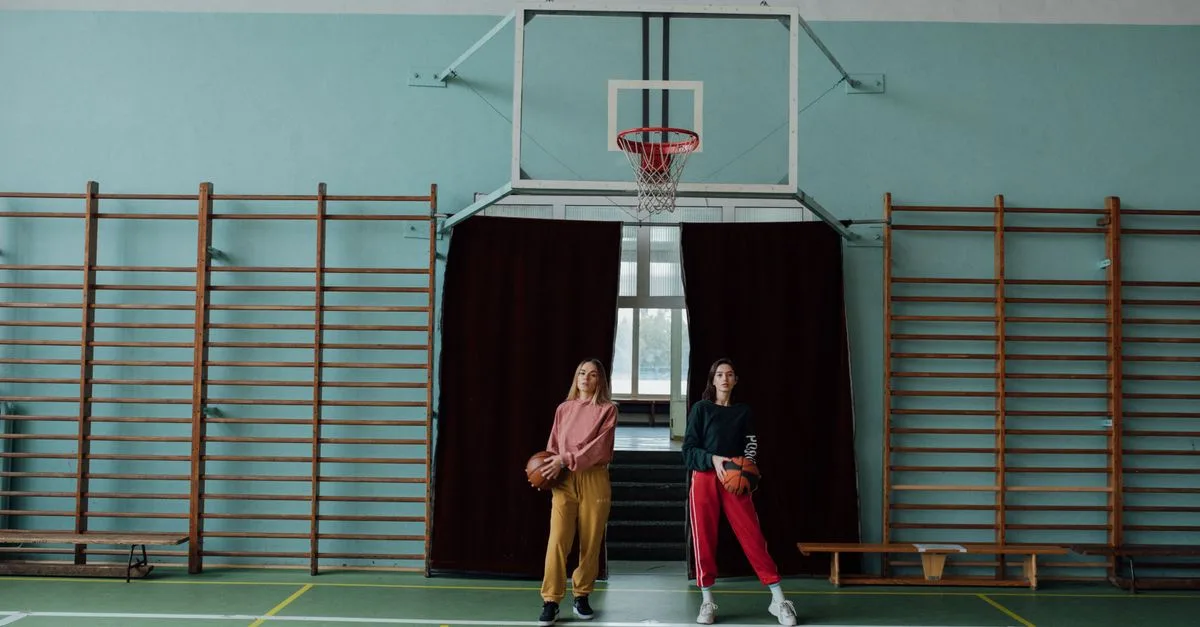 Two women holding basketballs in a gymnasium, standing by the hoop.