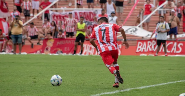 A soccer player in red and white stripes plays during a match, with fans watching from the stands.