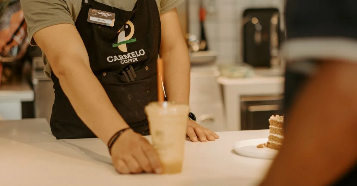 A barista wearing an apron serves an iced coffee to a customer inside a coffee shop.
