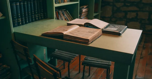 Warm-toned library interior featuring open books on a vintage table with green shelves.