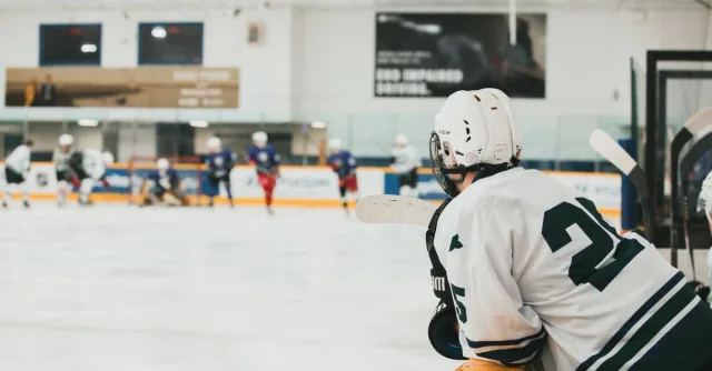 Adult ice hockey player in white jersey observing match from bench indoors.