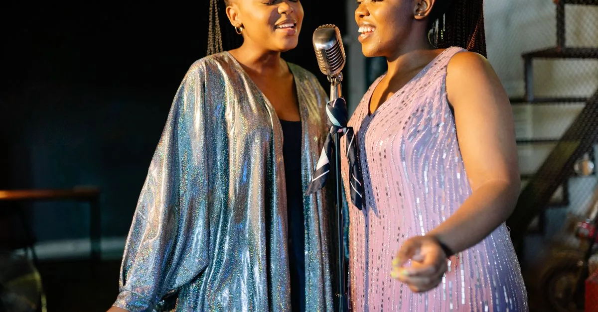 Two women joyfully singing with a vintage microphone on stage, dressed in glittery outfits.