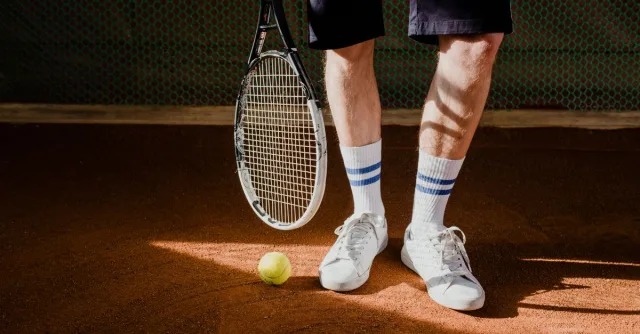 Close-up of a tennis player with racket and ball on a clay court, wearing white sneakers and socks.