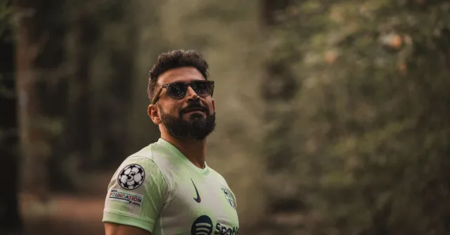 A bearded man in a soccer jersey stands confidently in a serene forest setting.
