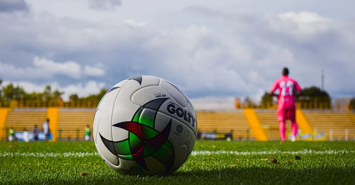 Close-up of a soccer ball on green grass field with a blurred player in pink jersey in the background during a match.