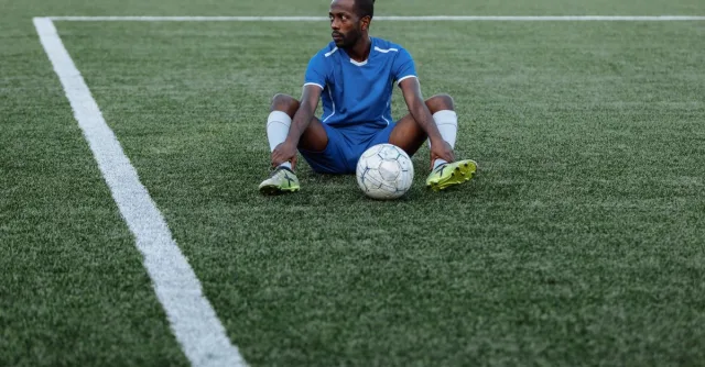 A soccer player in a blue uniform sits on a green field with a ball, reflecting focus and sportsmanship.